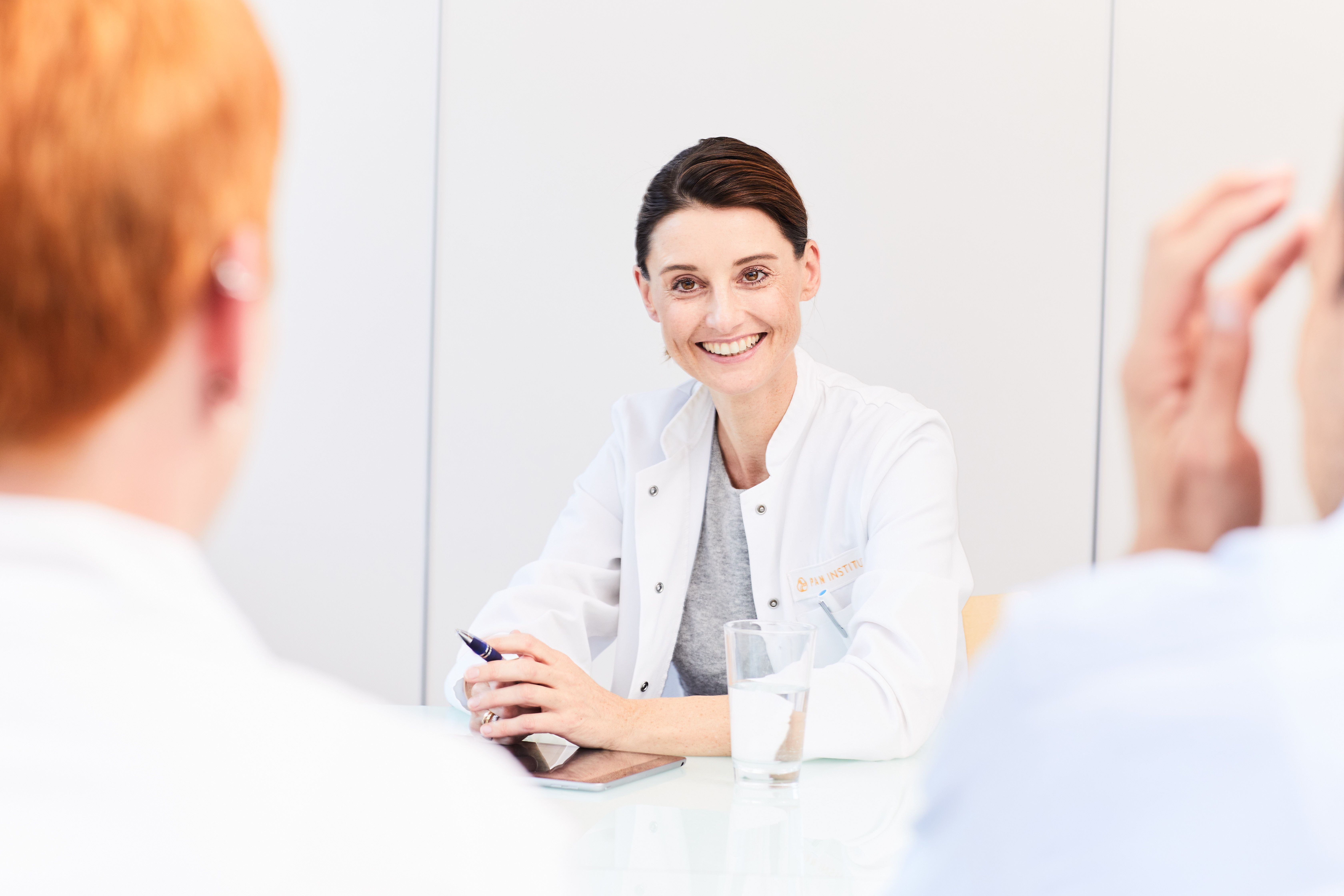 A smiling healthcare professional in a white coat sits at a table, engaging with two patients. The setting conveys a relaxed atmosphere, with a glass of water on the table, emphasizing a welcoming and friendly consultation.