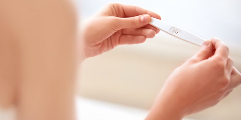 A close-up of two hands holding a pregnancy test stick, showing a positive result with two red lines. The background is softly blurred, indicating an indoor setting. The focus is on the test and the hands.