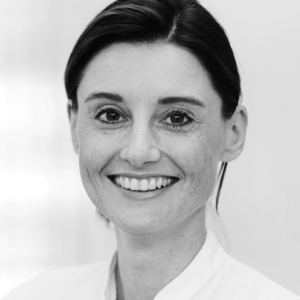 Eine lächelnde Frau mit kurzen, dunklen Haaren trägt ein weißes Oberteil. Sie schaut direkt in die Kamera und hat einen freundlichen Ausdruck. Der Hintergrund ist hell und unscharf.