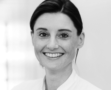 A smiling woman with medium-length, dark hair is wearing a white top. She has a friendly expression and is looking directly at the camera. The background is softly blurred, emphasizing her cheerful demeanor.