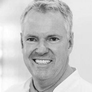 Ein lächelnder Mann mittleren Alters mit kurzen, grauen Haaren trägt einen weißen Kittel. Er steht vor einem hellen, unscharfen Hintergrund und wirkt freundlich und professionell.