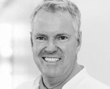 A smiling man with short gray hair is wearing a white coat. The background is bright and blurred, suggesting a professional setting, possibly a clinic or hospital. The man appears friendly and approachable.