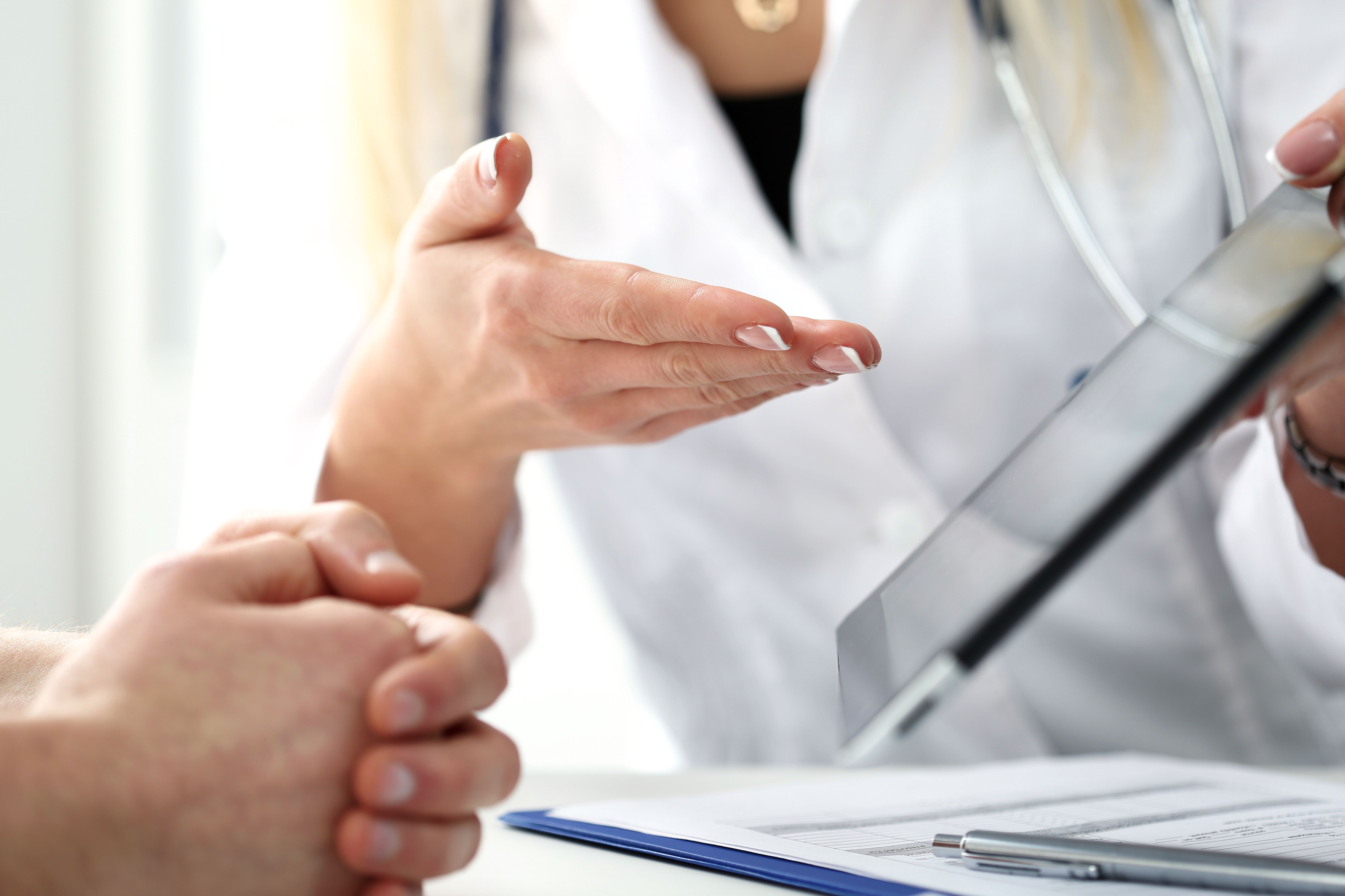 A healthcare professional gestures while discussing information on a tablet with a patient. The setting suggests a medical consultation, focusing on communication and patient care.