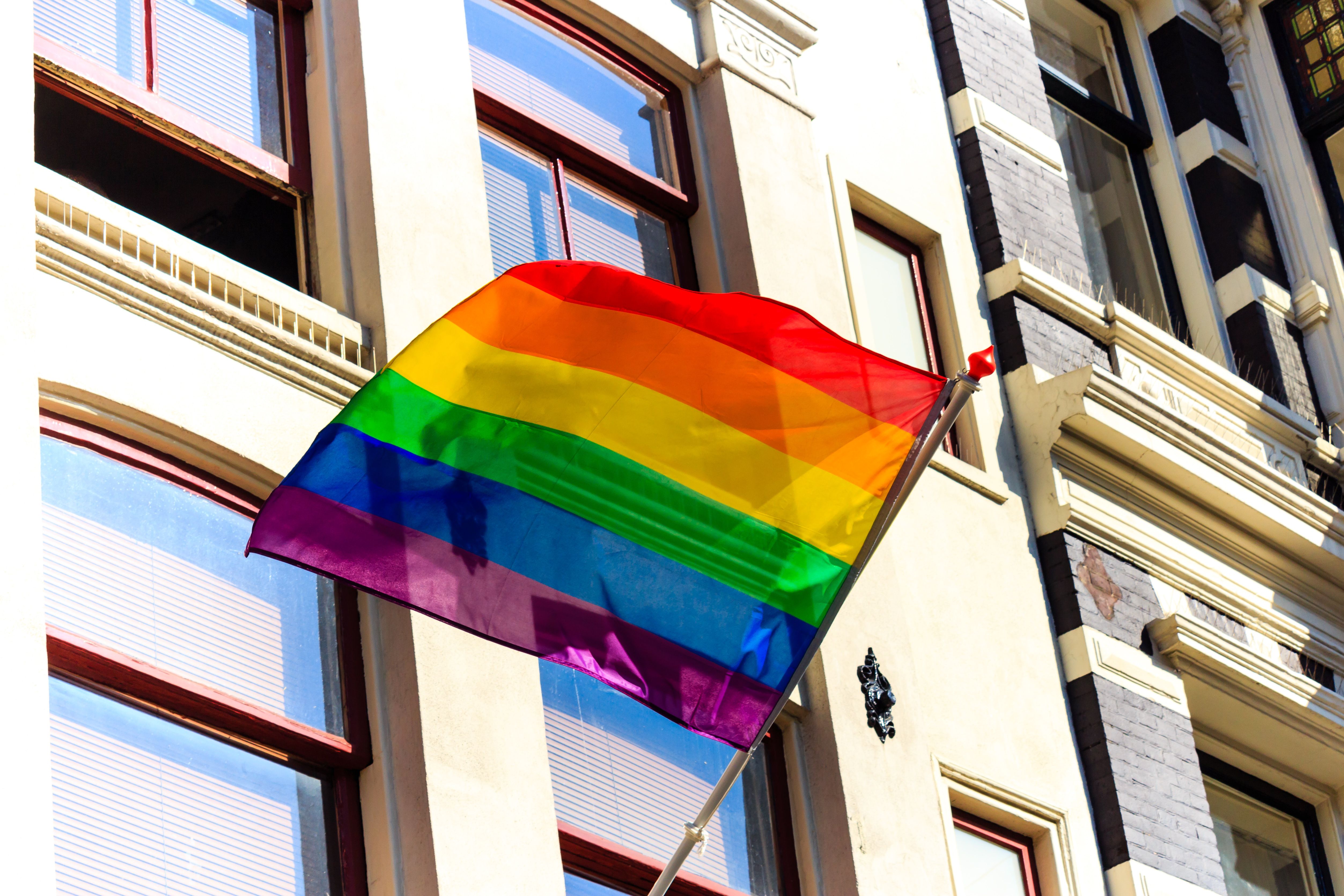 Eine Regenbogenflagge weht vor einem Gebäude. Die Flagge zeigt die Farben Rot, Orange, Gelb, Grün, Blau und Violett, die horizontal angeordnet sind und für Vielfalt und LGBTQ+-Rechte stehen.