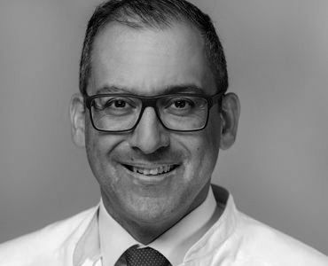 A smiling man wearing glasses and a white medical coat is depicted. He has short, dark hair and a friendly expression, conveying a professional and approachable demeanor. The image suggests a healthcare or medical context.