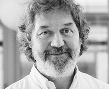 A middle-aged man with wavy hair and a beard smiles warmly at the camera. He wears a white shirt, and the background is softly blurred, suggesting an indoor setting. The image conveys a friendly and approachable demeanor.
