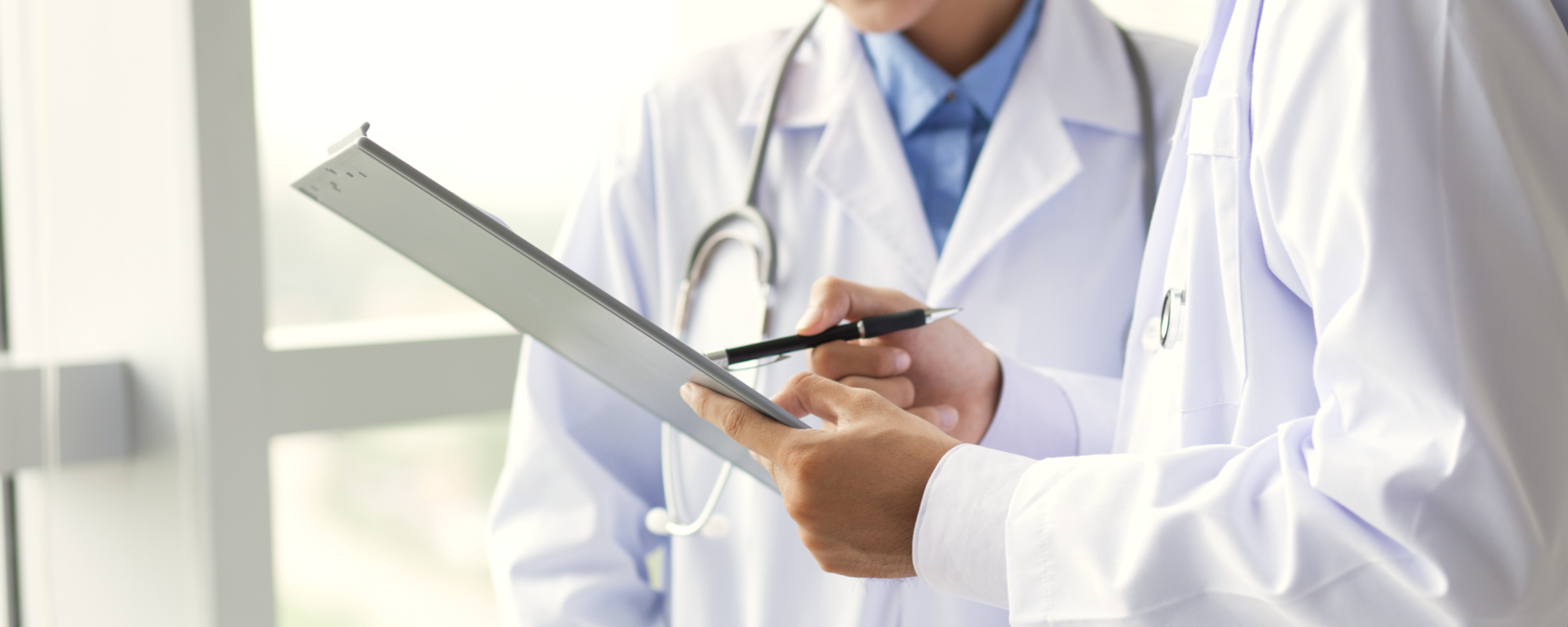 Two healthcare professionals in lab coats are discussing and reviewing information on a tablet. One is holding a pen, pointing at the screen, while the other is focused on the tablet, implying collaboration in a medical context. The setting appears well-lit and modern.