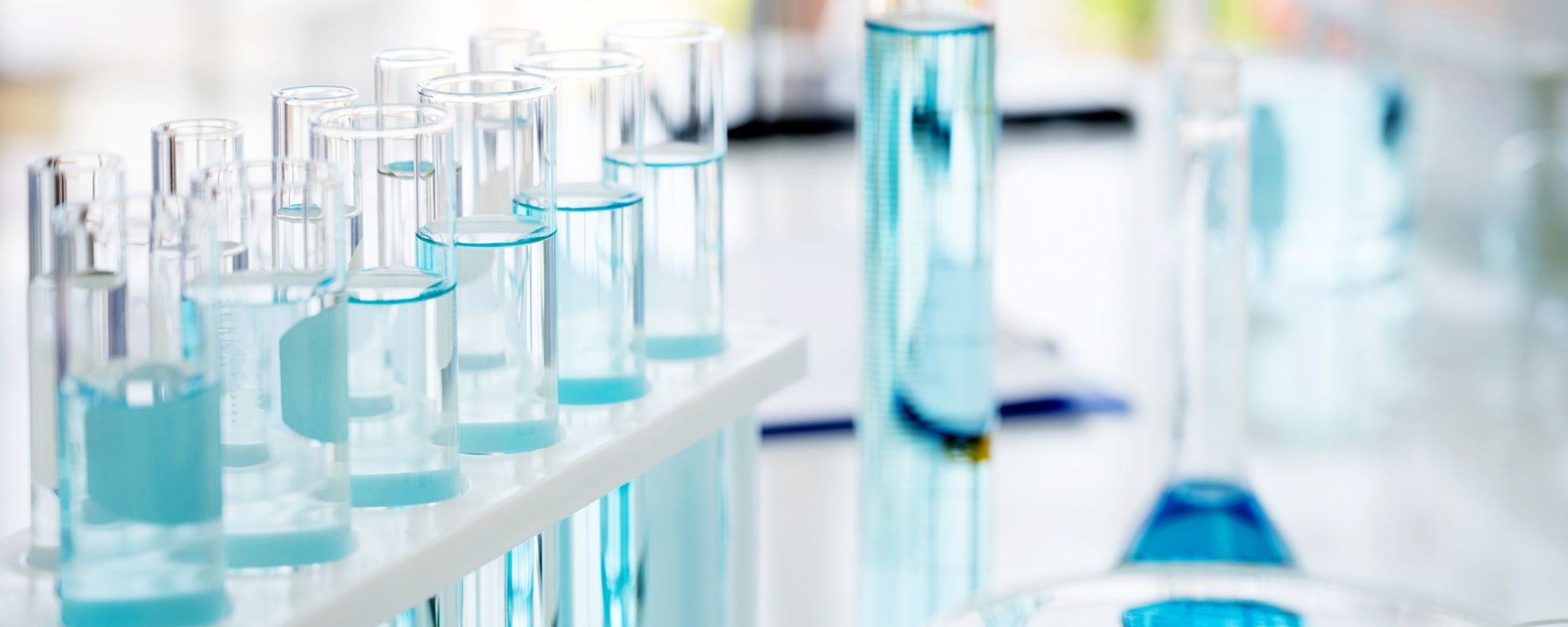 Auf dem Bild sind mehrere Reagenzgläser und eine Erlenmeyerkolben zu sehen, die mit blauen Flüssigkeiten gefüllt sind. Die Gläser stehen auf einem weißen Ständer und sind klar und sauber angeordnet, was eine wissenschaftliche Laborumgebung suggeriert.