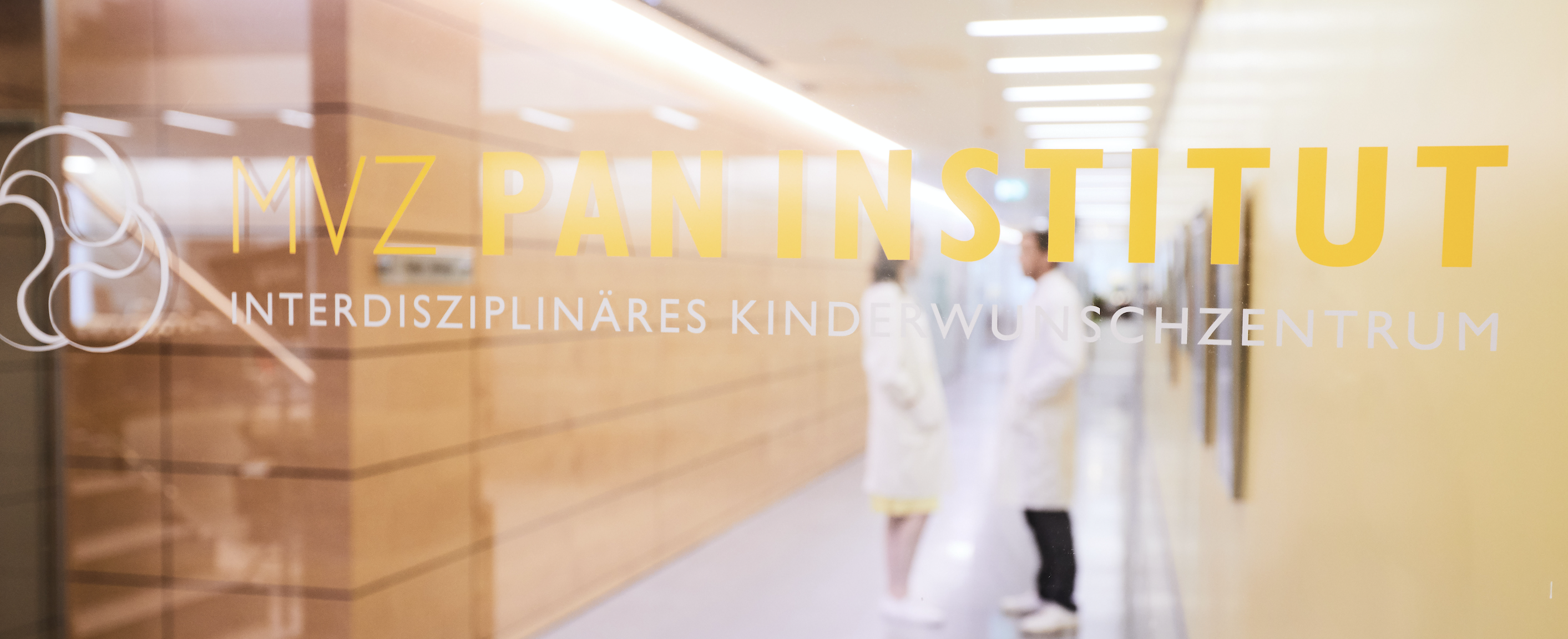 The image features a blurred interior corridor of a medical facility with two doctors in white coats conversing. Prominently displayed is a sign for "MVZ PAN INSTITUT - Interdisziplinäres Kinderwunschzentrum," indicating it is an interdisciplinary center focused on fertility and family planning.