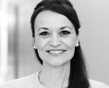 A smiling woman with long, dark hair styled to one side, wearing a light-colored top. The background is softly blurred, emphasizing her friendly expression. She appears confident and approachable, suitable for a professional profile.