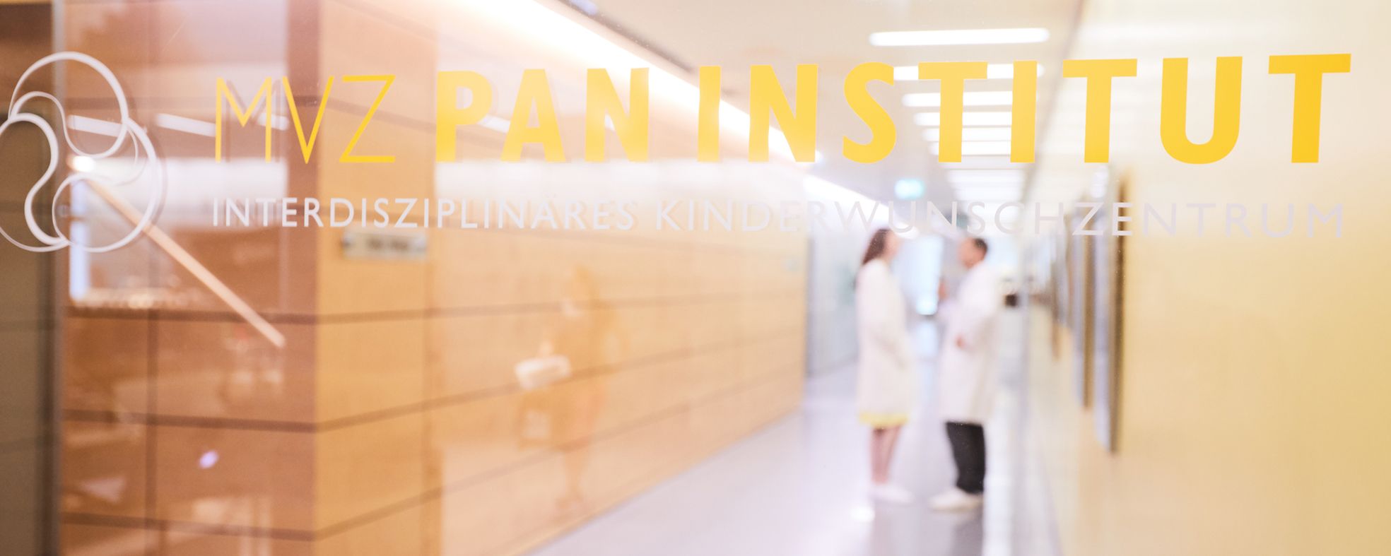 Das Bild zeigt den Flur des MVZ Pan Instituts, einem interdisziplinären Kinderwunschzentrum. Zwei Ärzte in weißen Kitteln stehen im Gespräch. Der Hintergrund ist unscharf, wobei Holzvertäfelungen und beleuchtete Wände sichtbar sind.