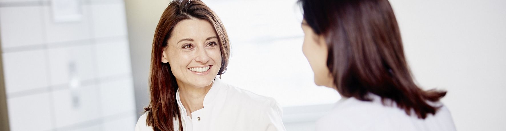 A healthcare professional in a white coat is engaged in a friendly conversation with a patient. The clinician is holding a tablet and smiling, suggesting a positive and informative discussion in a well-lit, modern setting.