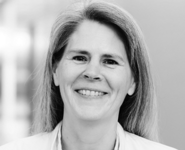 A woman with long, straight hair smiles warmly at the camera. She has a friendly expression and is wearing a light-colored top. The background is blurred, drawing attention to her face.