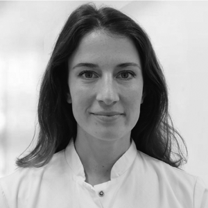 Porträt einer Frau mit langen Haaren, die einen weißen Kittel trägt. Sie lächelt leicht und blickt direkt in die Kamera. Der Hintergrund ist unscharf und hell.