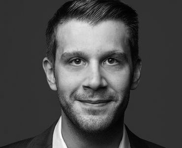 A smiling man with short hair and a slight beard is shown against a dark background. He is wearing a suit and a collared shirt, projecting a friendly and professional demeanor.