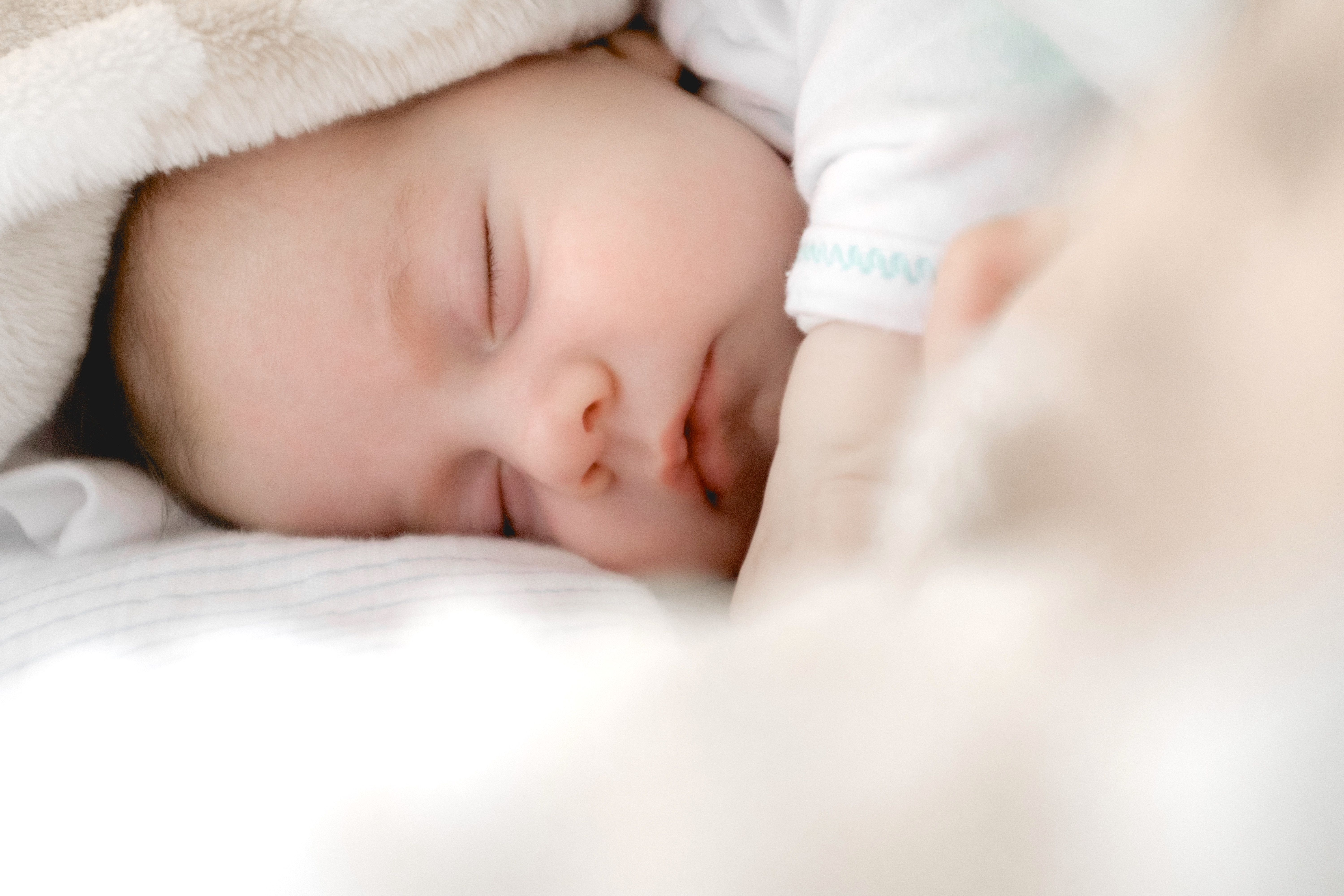 A peaceful sleeping baby is nestled under a soft blanket, with their eyes closed and a serene expression. Their small hand is gently resting, creating a tranquil and tender scene of innocence and comfort.
