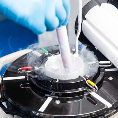 A gloved hand is holding a test tube above a black laboratory device that produces vapor. The device has multiple colored connectors and is likely used for sample processing in a scientific setting. The scene conveys a high-tech, controlled environment.