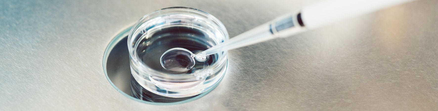 A close-up image shows a small glass dish containing a liquid, with a pipette hovering above it, ready to dispense the liquid. The surface beneath is metallic and smooth.