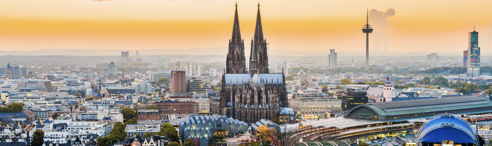Das Bild zeigt die ikonischen Türme des Kölner Doms silhouettiert gegen einen orangefarbenen Sonnenuntergang. Im Hintergrund sind weitere städtische Gebäude und der Fernsehturm von Köln zu sehen. Die Szene vermittelt ein Gefühl der Weite und Urbanität.