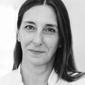 Ein Porträt einer Frau mit langen, braunen Haaren und einem freundlichen Ausdruck. Sie trägt ein weißes Oberteil und schaut direkt in die Kamera. Der Hintergrund ist unscharf und hell.