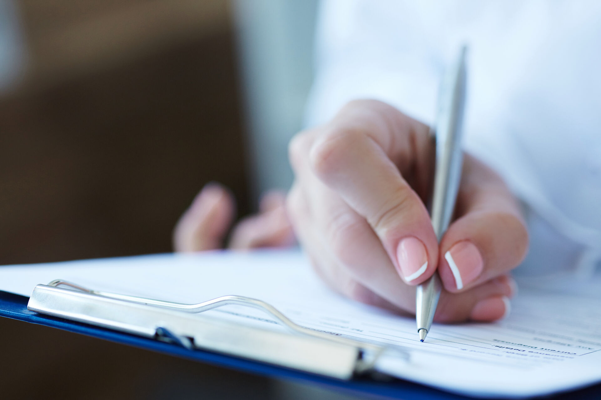 Eine Hand hält einen Stift und schreibt auf ein Dokument, das auf einem Clipboard liegt. Der Fokus liegt auf der präzisen Bewegung des Schreibens.