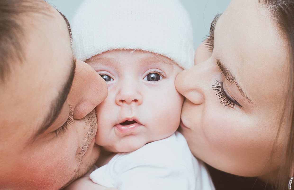 Ein Baby sitzt in der Mitte von zwei Elternteilen, die es sanft küssen. Das Baby trägt eine weiße Mütze und hat einen neugierigen Blick. Die Eltern zeigen Zuneigung und Liebe zueinander und zu ihrem Kind.