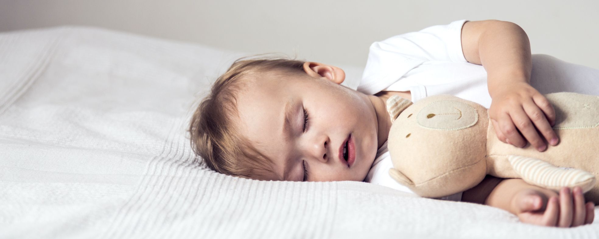 Ein schlafendes Kleinkind liegt auf einem Bett, umarmt ein plüschiges Spielzeug. Das Kind hat blondes Haar und trägt ein helles Oberteil. Die Umgebung ist einfach und der Hintergrund unauffällig, was eine ruhige und friedliche Atmosphäre vermittelt.