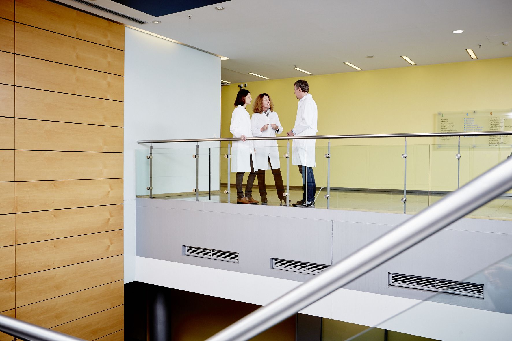 Three medical professionals in white lab coats stand on a balcony discussing together. They are positioned near a railing, and the setting features modern architecture with wooden elements and bright walls. The focus is on their conversation in a clinical environment.