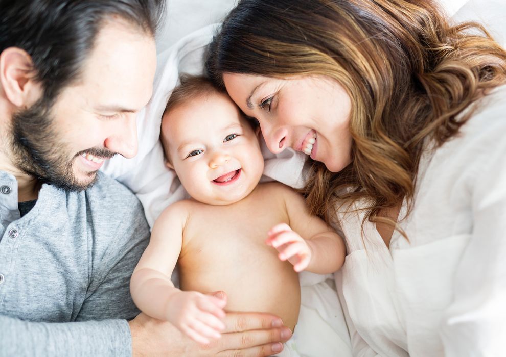 A smiling baby lies between a man and a woman, who are both smiling and looking at the baby. They are nestled together on a soft bed, creating a warm and loving family moment.