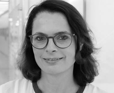 A woman with shoulder-length dark hair and glasses smiles gently at the camera. She is dressed in a light-colored top and appears confident and approachable, set against a neutral background.
