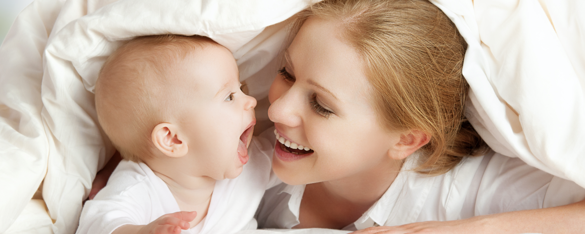A joyful woman smiles at an infant who is also smiling and opening their mouth as if to giggle or coo. They are covered by a soft blanket, creating a warm, intimate atmosphere that conveys love and happiness between them.