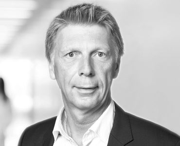 A middle-aged man with short, light-colored hair is looking directly at the camera. He is wearing a dark suit over a white shirt. The background is blurred, suggesting an indoor setting, possibly an office corridor. The image is in black and white.