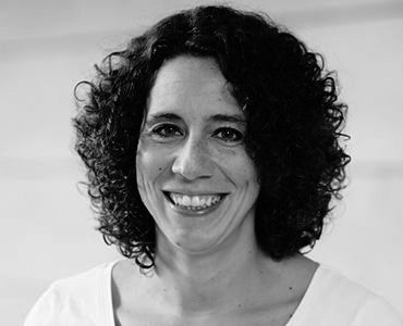 A smiling woman with curly hair is facing the camera. She is wearing a light-colored shirt and has a friendly expression. The image is primarily in black and white, focusing on her facial features and expression.