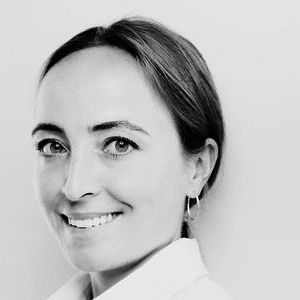 Eine lächelnde Frau mit langen, braunen Haaren und hellen Augen trägt ein weißes Hemd. Der Hintergrund ist schlicht und hell. Ihr Gesicht ist freundlich und offen, was eine positive Ausstrahlung vermittelt.