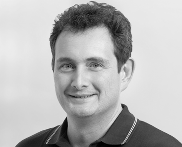 Portrait of a man with short, curly dark hair, smiling. He is wearing a black polo shirt with a white collar. The background is a soft, light color.