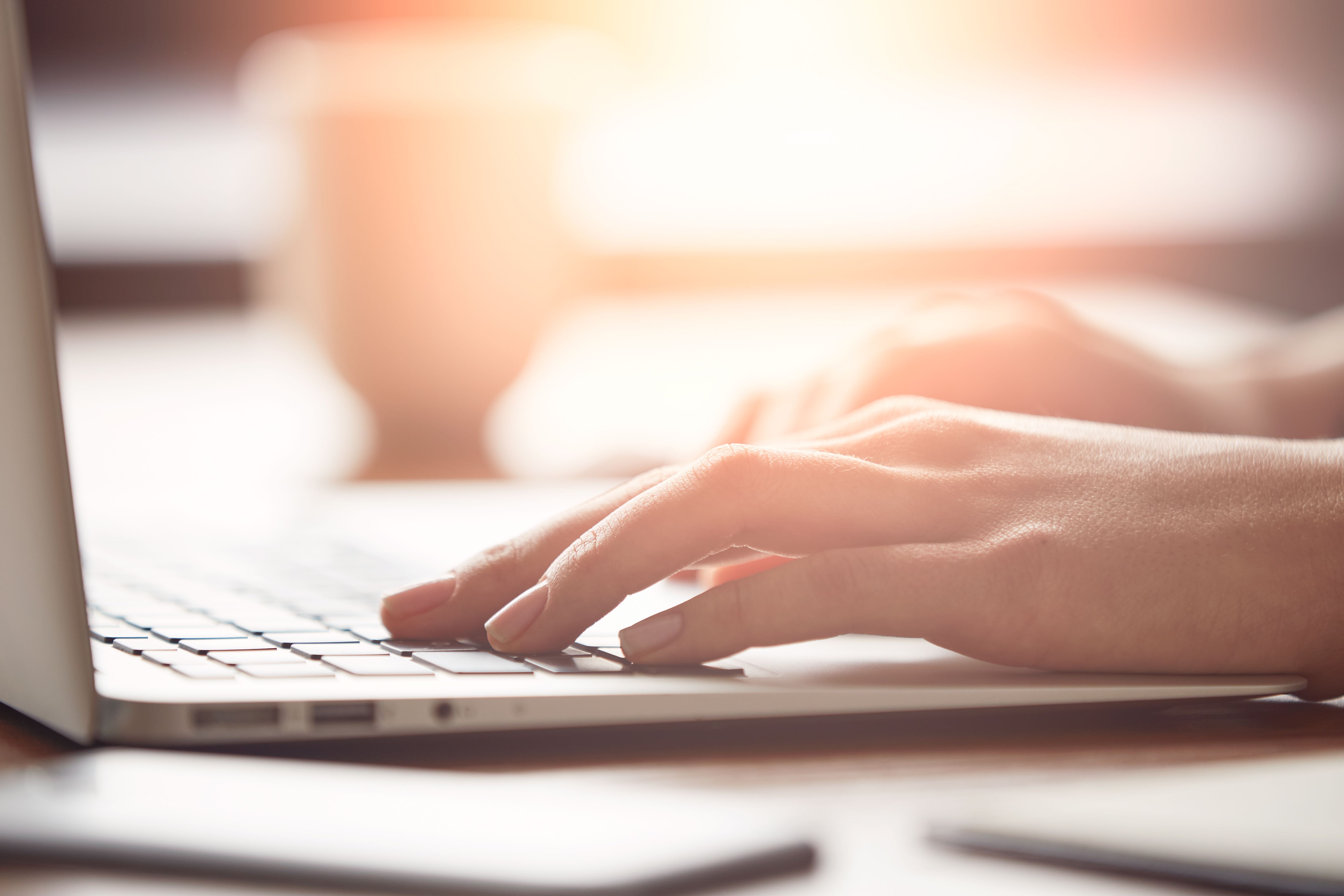 A close-up view of a person's hands typing on a laptop keyboard. The background is softly blurred, suggesting a warm, focused workspace environment. Light filters in, creating a bright atmosphere around the scene.