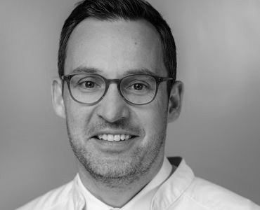 A smiling man with short, dark hair and glasses is wearing a white coat. He appears friendly and professional, likely in a medical or culinary setting. The background is a soft, neutral color, enhancing his approachable demeanor.
