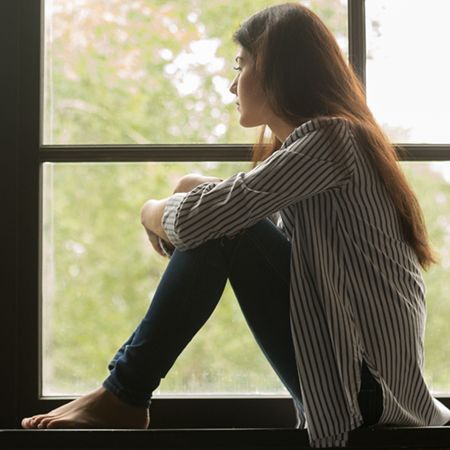 A young woman sits by a window, gazing outside with a pensive expression. She is barefoot, wearing a striped shirt and jeans, and appears to be deep in thought, surrounded by soft natural light and greenery in the background.
