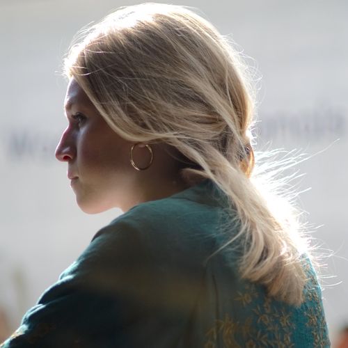 A profile view of a woman with long, light hair styled in loose waves. She wears hoop earrings and a patterned teal top, with soft sunlight highlighting her features against a blurred background of people in casual attire.