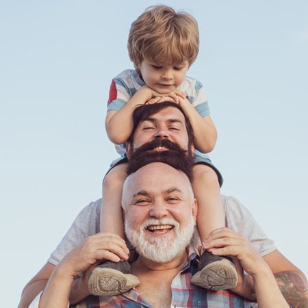 A joyful family moment featuring three males: a smiling older man at the front, a man with a beard in the middle, and a young boy sitting on the man's shoulders. They are outdoors against a clear sky, all appearing happy and engaged with each other.