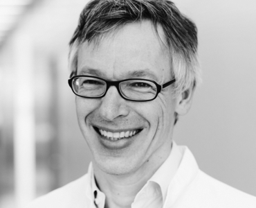 A middle-aged man with glasses and gray hair smiles warmly. He is wearing a white coat, suggesting he is in a medical or scientific setting. The background is softly blurred, enhancing the focus on his friendly expression.
