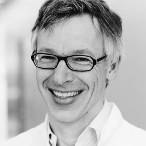 Ein lächelnder Mann mit Brille trägt einen weißen Laborkittel. Der Hintergrund ist unscharf und hell, was auf einen klinischen oder wissenschaftlichen Raum hindeutet. Sein freundlicher Gesichtsausdruck vermittelt Professionalität und Zugänglichkeit.