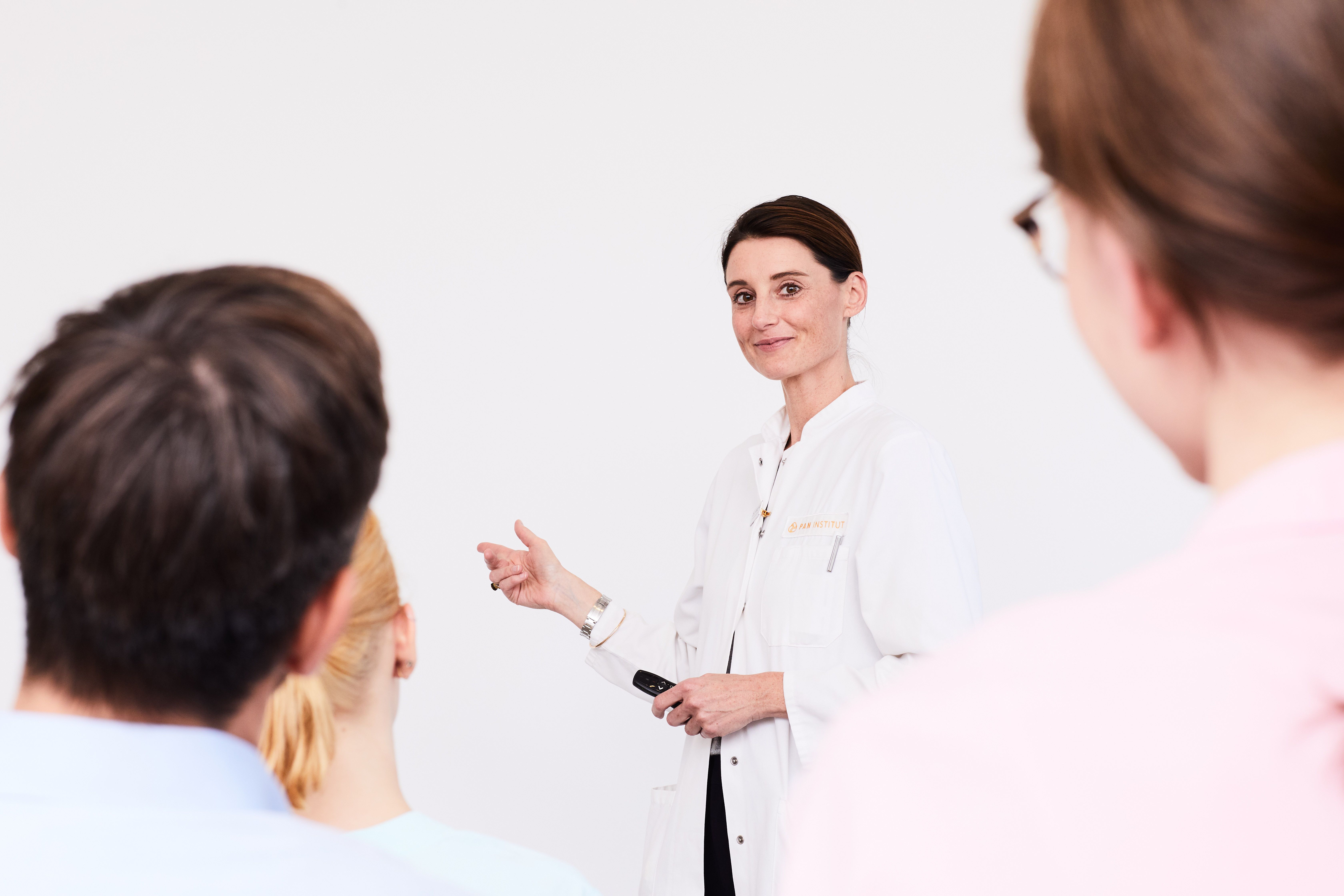 A doctor in a white coat stands confidently, addressing an audience. She gestures with her hand, holding a pen or pointer, while the backs of several heads are visible, indicating that she is presenting or teaching a group.