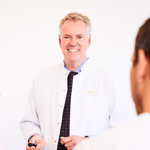 A smiling male doctor in a white coat stands in front of a small audience, presenting information. The audience consists of three people, who are facing him and listening attentively. The background is plain and bright, focusing on the speaker and his engaging demeanor.
