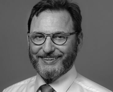 A middle-aged man with a beard and glasses smiles warmly at the camera. He is wearing a light-colored dress shirt and a tie. The background is plain and neutral, focusing attention on his friendly expression.
