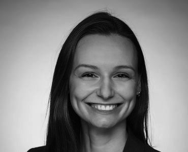 A woman with long, dark hair smiles broadly at the camera. She exudes a friendly and confident demeanor. The image is in black and white, highlighting her features against a neutral background.