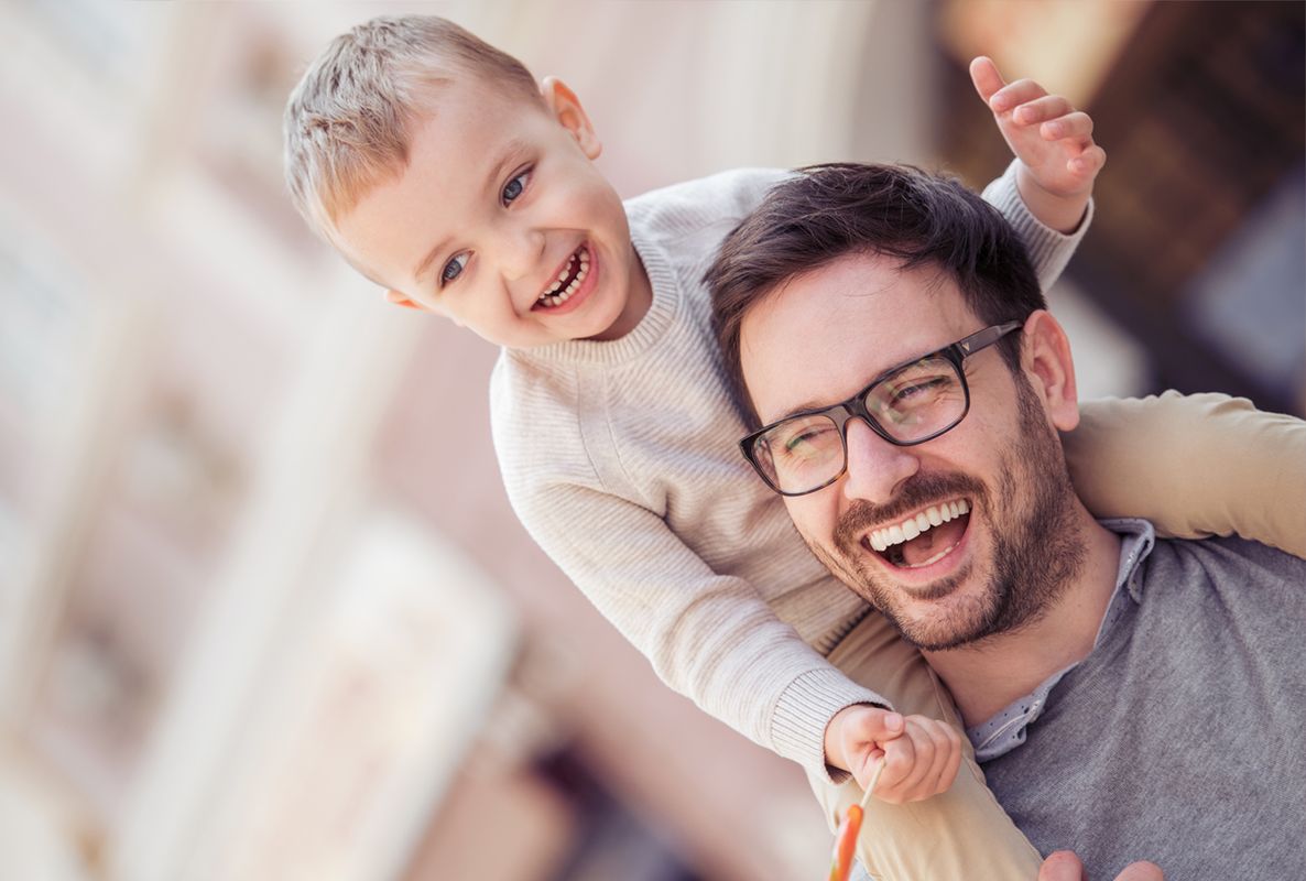 Ein lächelnder Vater trägt seinen fröhlichen Sohn auf den Schultern. Beide lachen und scheinen eine unbeschwerte Zeit miteinander zu verbringen. Der Hintergrund vermittelt eine freundliche Atmosphäre.