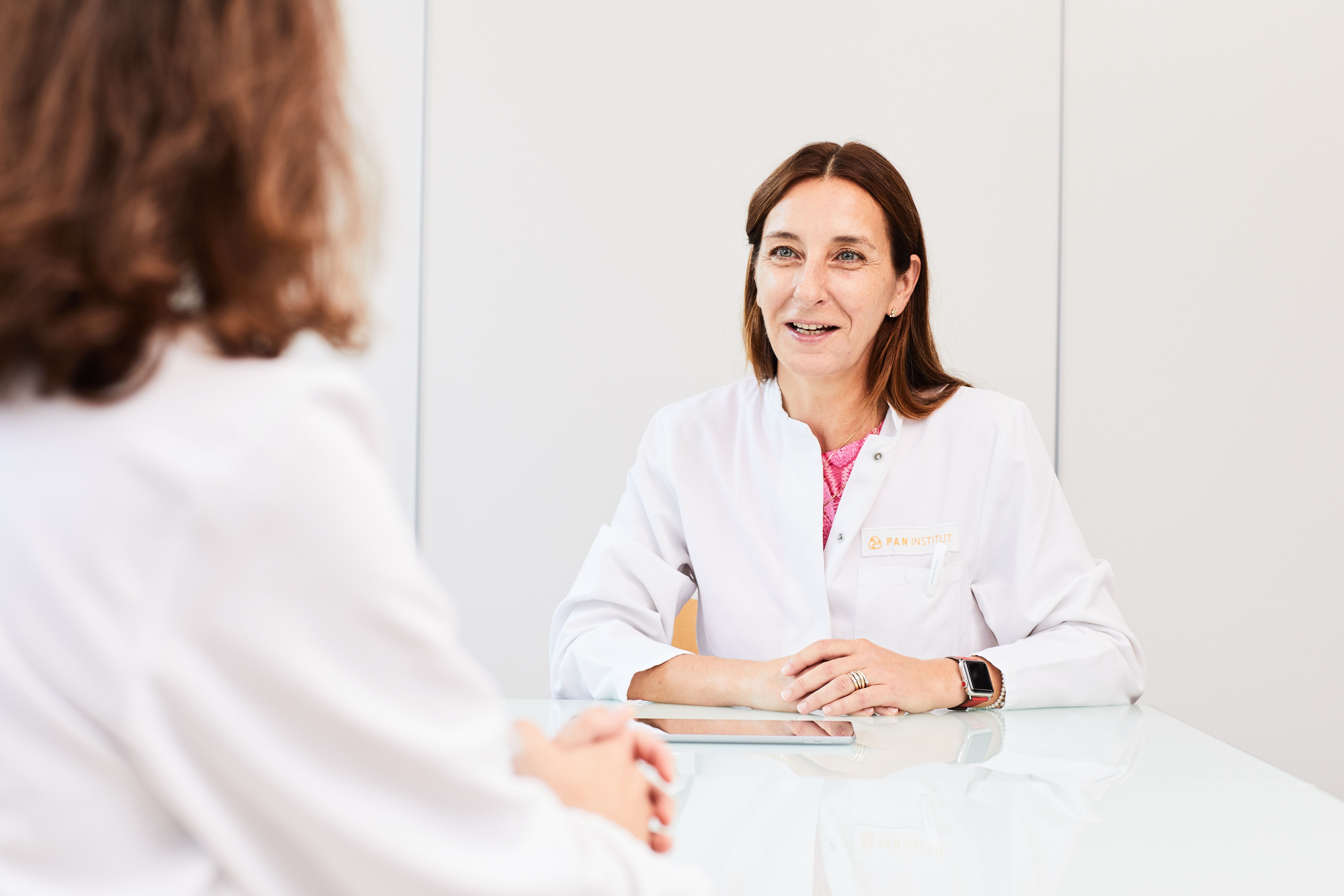 A healthcare professional in a white coat is sitting at a table, smiling and engaging in conversation. The focus is on the interaction between her and a person whose back is facing the viewer. The setting appears to be a bright, modern consultation room.