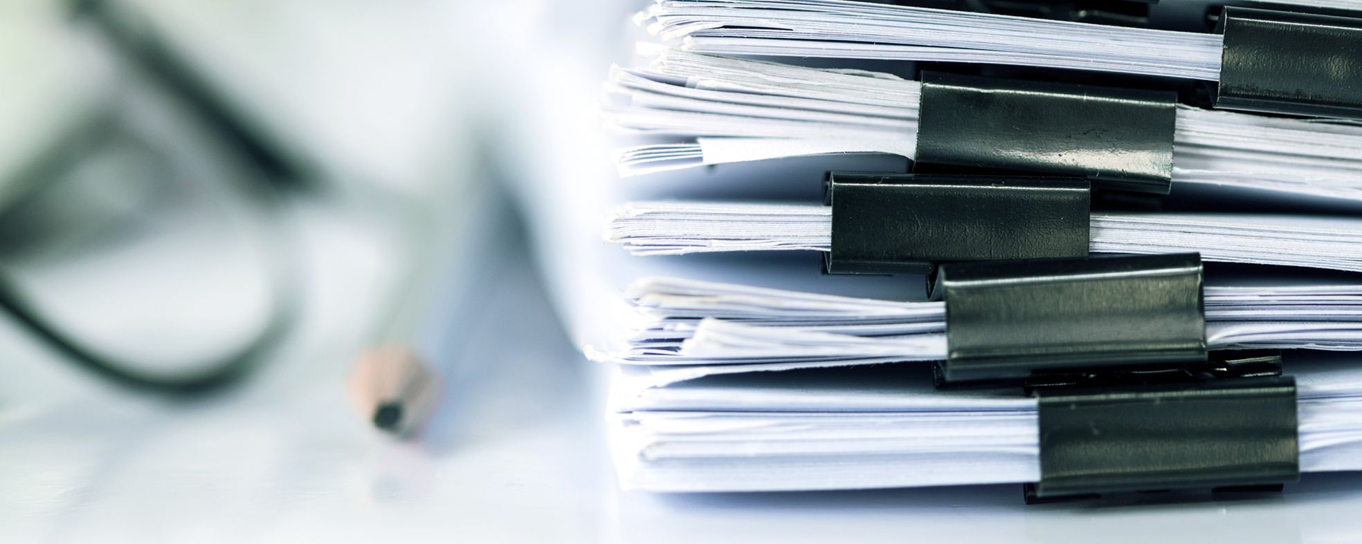 A close-up image of stacked documents secured with black binder clips, set against a blurred background that includes a pen. The focus is on the organized papers, suggesting a theme of paperwork or office work.