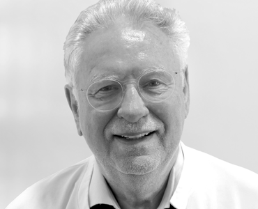A smiling older man with glasses and light gray hair appears in the image. He is wearing a white coat, suggesting he is a healthcare professional. The background is light and neutral, focusing on his friendly demeanor.